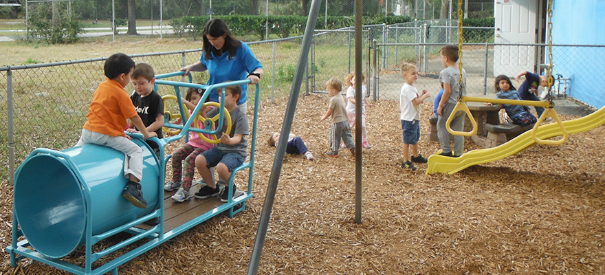 Children and a teacher using playground equipment in a fenced outdoor play area.